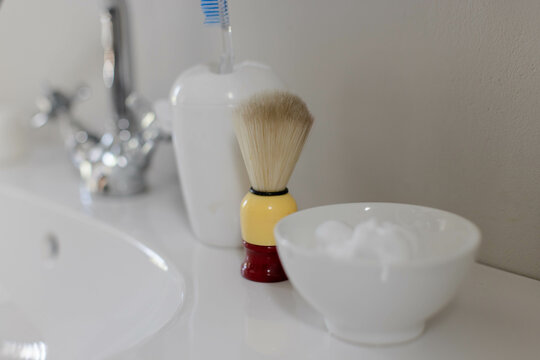 Shaving brush standing on bathroom vanity beside white sink, toothbrush holder and foam bowl