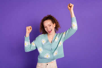 Cheerful young woman in a floral cardigan celebrates with raised arms against a vibrant purple...