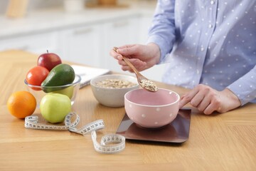 Woman weighting oatmeal on kitchen scale at wooden table indoors, closeup