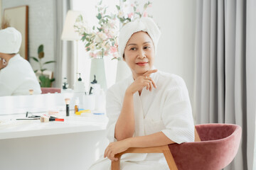 Asian senior woman sitting in beauty corner wearing spa robe and towel on head looking relaxed and elegant in front of vanity with cosmetics expressing confidence wellness aging grace self care pride
