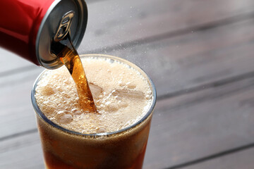 Pouring refreshing cola from can into glass at wooden table, closeup. Space for text