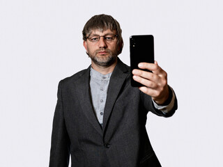 Men in grey suit holding phone in his hand, white background. Male model in his 50s with grey beard. Serious face expression. Video call concept.