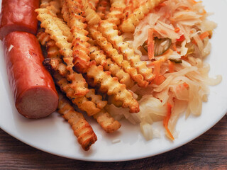 A plate of food with a hot dog and French fries on it and sauerkraut salad. The plate is on a wooden table. Tasty dinner.