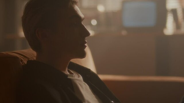 Young reflective man sitting on couch in living room filled with hazy warm light, looking thoughtfully ahead. Side view, tilt up shot