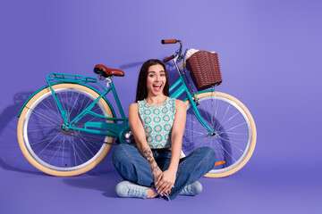 Smiling young woman sitting near a vintage bicycle in stylish casual outfit against a purple...