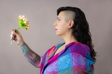 Serious woman in profile holding a small bouquet of flowers. She wears a colorful shawl, eyebrow piercing, and has a modern half shaved hairstyle. Horizontal studio portrait with gray background.
