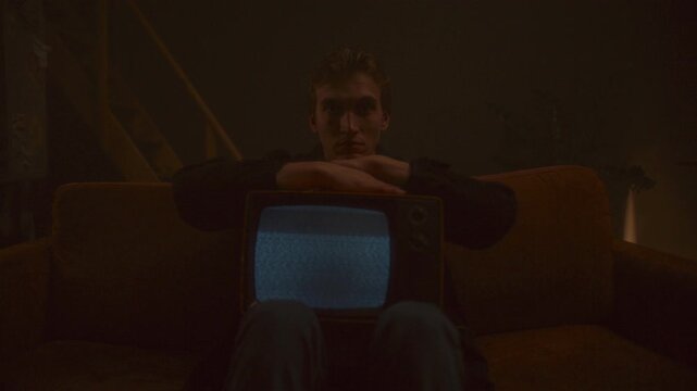 Young man sitting on couch with arms resting on retro TV showing static screen in dimly lit room. Zoom shot, video portrait