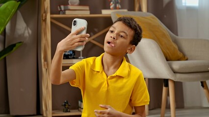 Young african american child boy spending free time at home, kid sitting on the floor in the living room and taking funny selfies on smartphone.