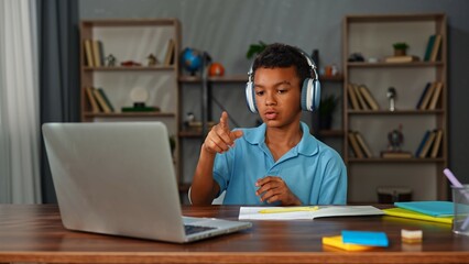 Young african american child boy studying at home, kid sits at desk attends school class online on laptop, wearing headphones and talking with teacher, answers question.