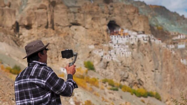 Rack focus shot of an Indian male influencer shooting video of Phugtal monastery with his mobile phone on a gimbal at Zanskar valley in Ladakh, India. Influencer shooting content for social media.
