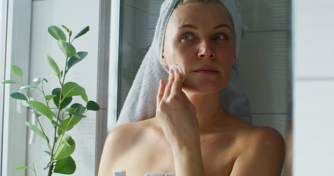 A woman takes care of her face, she wipes her face with a cotton pad while looking in the mirror. Close-up of her reflection in the mirror. Bathroom at home