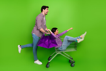 Excited young couple having fun with shopping cart and festive bags on vibrant green background, enjoying the shopping experience