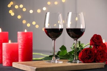 Glasses of red wine, burning candles and roses on table against gray background with blurred lights. Romantic dinner