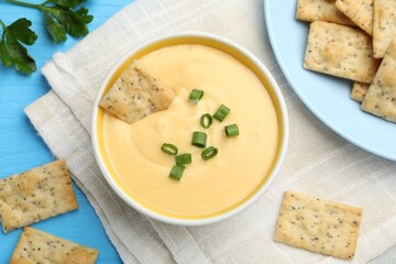 Tasty cheese dipping sauce in bowl and crackers on light blue wooden table, flat lay