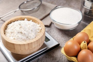 Digital kitchen scale with bowl of flour and other products on grey textured table, closeup