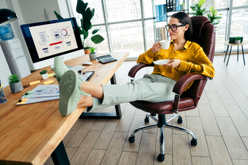 Young businesswoman relaxing at her desk in a modern office during daylight enjoying a coffee...