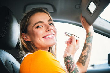 Young woman applying lipstick in a car while smiling, showcasing style and beauty during daylight...