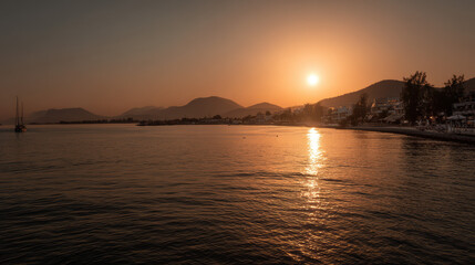 A tranquil sunset paints the water with golden hues as a lone sailboat glides near the horizon, framed by distant mountains and a serene coastal town glow.