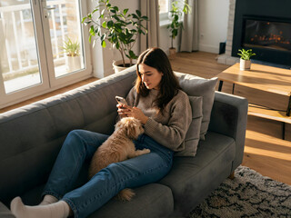 Naklejka na ściany i meble Young freelance woman sitting on couch sofa using mobile phone application, chatting online. Relaxing girl at real estate apartment home chill out holding gadget at sunny day petting pet dog