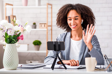 Client consultation on style and design. Smiling african american woman with wireless headphones waving to webcam in interior of fashion studio, free space