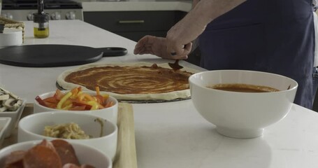 Man evenly spreading sauce on pizza dough with spoon on kitchen counter - medium, tracking shot