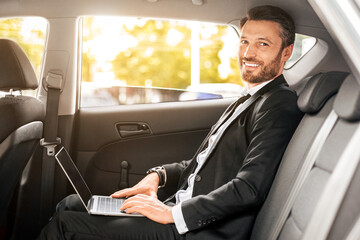 Cheerful businessman sitting in car, using laptop, working online. Handsome man in suit working on...