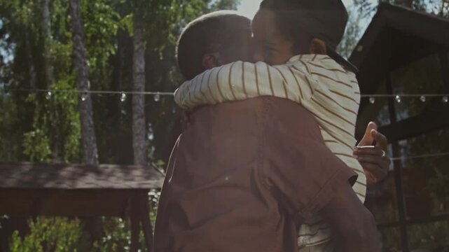 Slow motion medium closeup of proud African American father lifting up and kissing baby son, while playing together outdoors on hot summer day, in bright sunlight with strong lens flare