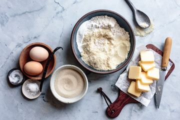 An assortment of baking ingredients including eggs, flour, sugar, butter, and vanilla beans is arranged on a marble surface.