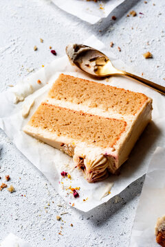 A piece of two-layer vanilla cake with frosting on parchment paper, accompanied by a gold spoon.