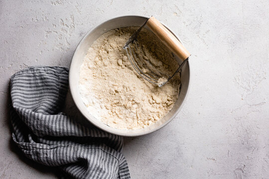 A bowl of flour mixture with a pastry cutter on a textured surface, accompanied by a striped kitchen towel.