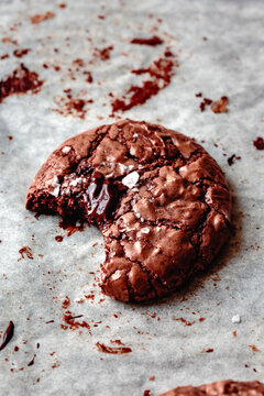 A close-up of a chocolate cookie with a bite taken out, showing gooey chocolate inside and sprinkled sea salt on top, resting on parchment paper.