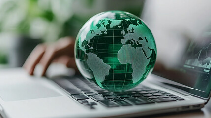 Green countries on a transparent glass globe rest over a blurred laptop keyboard displaying financial graphs, symbolizing global business, sustainable investment, and eco-conscious finance.

