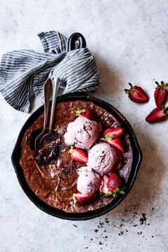 A skillet brownie topped with scoops of strawberry ice cream and fresh strawberries, served with two spoons on a textured surface.