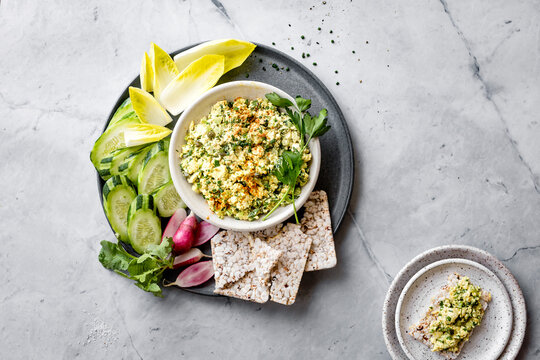 A bowl of creamy dip with herbs is served alongside sliced cucumbers, endive leaves, radishes, and rice cakes on a marble surface.