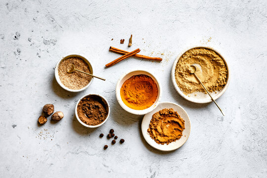 An assortment of spices in ceramic bowls and plates on a textured surface, including cinnamon sticks, nutmeg, and various ground powders.