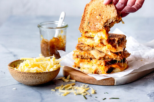 A hand reaches for a stack of grilled cheese sandwiches on a wooden board, with a bowl of grated cheese and a jar of chutney nearby.