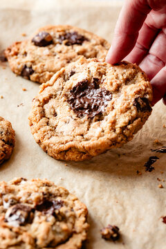 A hand holding a freshly baked chocolate chip cookie on a parchment-lined baking sheet.