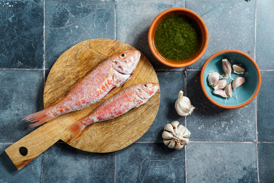 resh red mullets on a rustic wooden cutting board with olive oil, garlic, and green sauce on a blue tile background