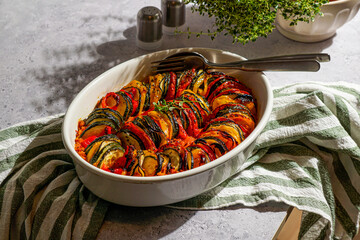 beautifully arranged ratatouille, featuring sliced zucchini, eggplant, tomatoes, and yellow peppers garnished with fresh herbs. Warm sunlight enhances the cozy atmosphere