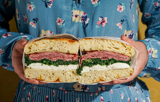 woman in blue dress holding a focaccia bread italian sandwich, Porchetta meat, kale, mozzarella, pesto, sun light modern