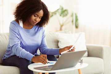 Portrait of professional black woman at work, reading and writing some papers sitting on couch at table with pc