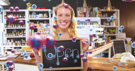 Barista holding open sign in artisanal cafe, with counter and pastries, coronavirus graphics