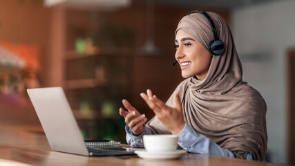 Fototapeta premium Young woman in hijab having video conference at cafe, using headset and laptop, drinking coffee, empty space