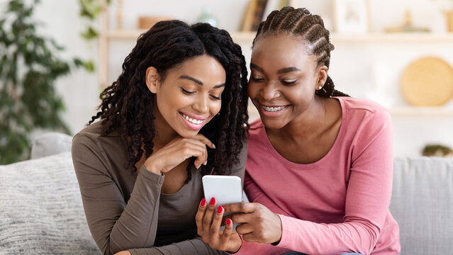Smiling african american girlfriends checking photos on mobile phone, spending time together at home, copy space. Two young black ladies sitting on sofa at living room, using modern smartphone