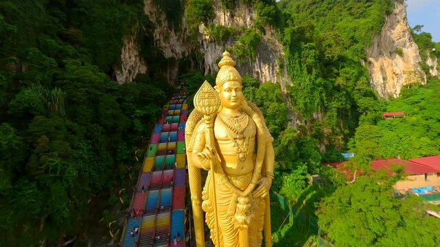 FPV drone shot of Batu Caves and golden Lord Murugan statue in Kuala Lumpur, Malaysia. Famous religious landmark with colorful stairs, limestone cliffs, and lush greenery.