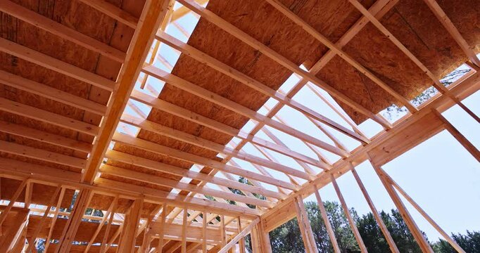 Wooden roof trusses and framing during building construction, showing structural framework with oriented strand board and exposed wooden beams, with trees visible in background.