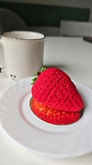 Strawberry-shaped mousse dessert on a white plate with a coffee mug in the background. This image...
