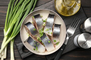 Pieces of delicious herring with green onions served on black wooden table, flat lay