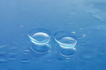 Pair of contact lenses and water drops on blue mirror surface, closeup