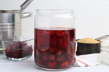 Tasty cherry compot in glass jar and ingredients on white tiled table, closeup